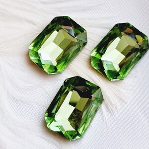 May include: Three faceted, rectangular green gemstones with a light green hue. The gems are arranged on a white, textured surface, possibly fabric or feathers. The gemstones have a polished, reflective surface.
