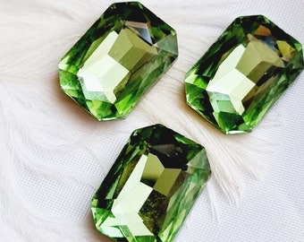 May include: Three faceted, rectangular green gemstones with a light green hue. The gems are arranged on a white, textured surface, possibly fabric or feathers. The gemstones have a polished, reflective surface.