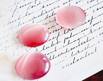 May include: Three pink glass cabochons with a white iridescent sheen. The cabochons are round and smooth, and they are arranged in a row on a piece of white paper with black handwriting.