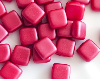 May include: A pile of small, square, red beads. The beads have a matte finish and are scattered on a white surface.