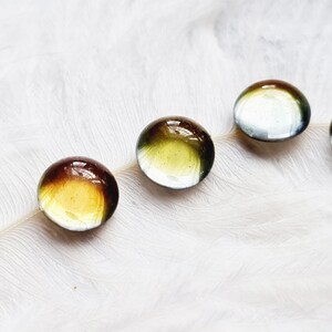 Puede incluir: Cuatro canicas de vidrio redondas con un esquema de color verde, marrón y amarillo en capas. Las canicas están dispuestas en fila sobre un fondo de pluma blanca.