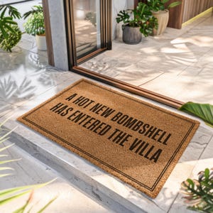 May include: A rectangular tan coir doormat with a black double-line border. The text A HOT NEW BOMBSHELL HAS ENTERED THE VILLA is printed in bold black capital letters across the center of the mat, positioned on a light-colored tiled entryway floor.