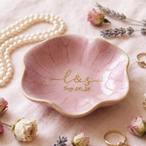 Pode incluir: Um prato de cerâmica rosa com borda ondulada, com letras douradas que dizem "L&S" e "Sep.06.26". O prato é rodeado por um colar de pérolas, anéis de ouro, rosas secas e raminhos de lavanda, tudo disposto sobre um tecido rosa.