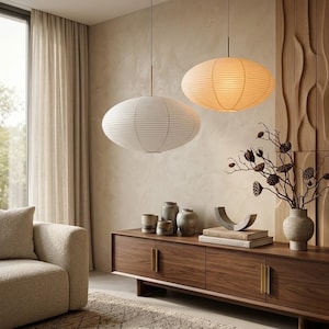 May include: Two oval-shaped pendant lights with ribbed paper shades hang from the ceiling. One light is white and the other is a warm beige. They are suspended by thin metal rods above a wooden sideboard featuring brass handles and ceramic vases.