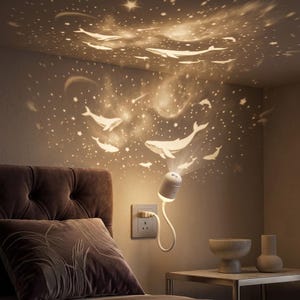 Puede incluir: Una lámpara proyector de pared blanca con cuello flexible que proyecta un patrón de luz cálida con formas de ballenas, estrellas y figuras celestiales sobre la pared y el techo encima de un cabecero capitoné marrón oscuro.
