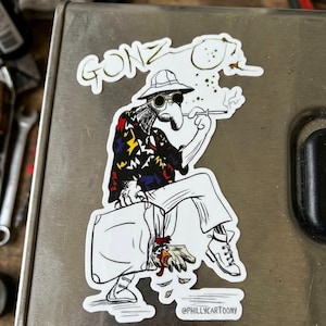 May include: A sticker featuring a cartoon illustration of a figure in a hat and sunglasses smoking a cigarette. The figure is wearing a patterned shirt and sitting on a suitcase with a chicken. The word "GONZ" is at the top. The artist's name, "@PHILLYCARTOONY", is at the bottom.