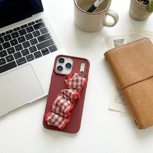May include: A dark red smartphone case featuring a built-in, ruffled fabric strap with a red and white checkered pattern. The case has a rectangular camera cutout and is positioned on a white surface next to a laptop, a mug, and a notebook.