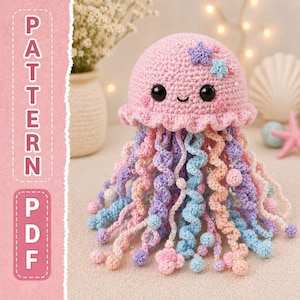 May include: A crocheted jellyfish toy in shades of pink, blue, purple, and peach. The jellyfish has a smiling face, star accents, and long, curly tentacles. The image also includes the text "PATTERN PDF" in a pink box.