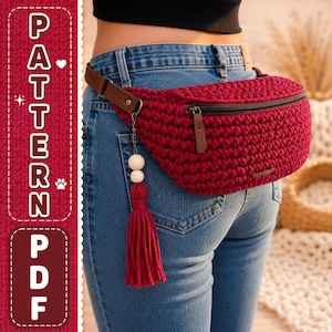 Peut inclure: Un sac banane en crochet rouge foncé avec une sangle en cuir marron foncé et un pompon en fil rouge assorti orné de deux perles en bois clair. Le sac possède une fermeture éclair horizontale et se porte à la taille sur un jean bleu.