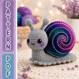 May include: A gray crocheted snail toy with a spiral shell featuring a gradient of blue, purple, and magenta yarn with silver metallic accents. The snail has black safety eyes, small purple cheeks, and two antennae. The text PATTERN PDF is visible.