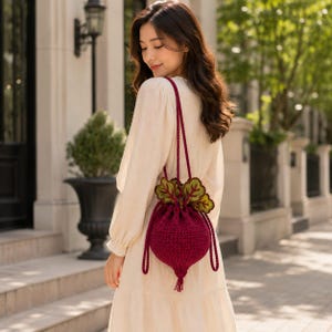 May include: A deep burgundy crochet drawstring bag shaped like a beetroot, featuring two long thin straps and decorative green and yellow crocheted leaves at the top. The bag has a textured yarn construction with a small tassel at the bottom point.