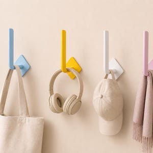 Peut inclure: Quatre crochets muraux de couleurs bleu, jaune, blanc et rose, chacun supportant un objet différent : un sac fourre-tout, un casque audio, une casquette de baseball et une écharpe. Les crochets sont fixés sur un mur beige clair.