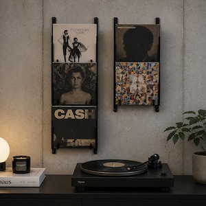 Könnte beinhalten: Ein schwarzer Plattenspieler mit einer Schallplatte steht auf einer schwarzen Oberfläche. Zwei schwarze, an der Wand befestigte Schallplattenhalter zeigen Albumcover. Eine kleine Topfpflanze und eine Tischlampe mit weißem Kugelschirm sind ebenfalls zu sehen.