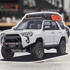 May include: A white Toyota 4Runner off-road vehicle sticker featuring a black front bumper, a roof rack with an orange traction board, and a light bar. The front skid plate displays the text CA FABRICATIONS. The vehicle has large tires.