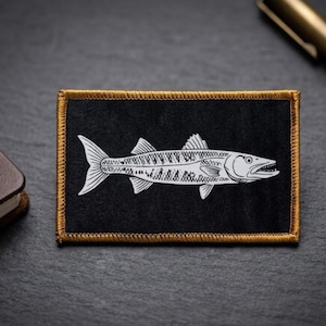 May include: A rectangular patch featuring a black fabric background with a white line art illustration of a barracuda fish in the center. The patch is finished with a thick, metallic gold embroidered border that frames the entire rectangular shape.