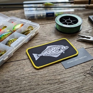 May include: A rectangular black patch with a yellow border featuring a white line-art illustration of a flatfish. Next to the patch is a small black card with the text OCEAN CREST PATCHES, fishing lures in a clear plastic box, pliers, and line.