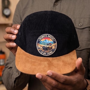 May include: Black corduroy cap with a brown suede brim. The cap features a circular patch with a mountain scene and the text "REMOTE AREAS IS MY THING". A stylish accessory for outdoor enthusiasts.