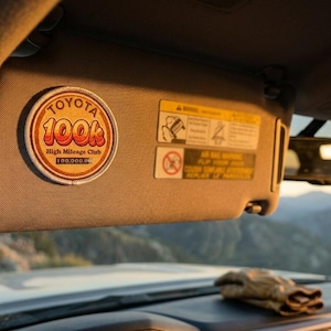May include: A circular embroidered patch attached to a gray vehicle sun visor. The patch features a white border, a tan background, and orange text that reads Toyota 100k High Mileage Club 100,000 miles. The visor also displays two rectangular safety labels.