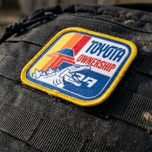 May include: A black tactical backpack featuring a rectangular embroidered patch with a yellow border. The patch displays a white dinosaur graphic, a red and orange striped logo, and the text TOYOTA OWNERSHIP in blue and red lettering.