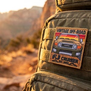 May include: A rectangular embroidered patch attached to an olive green tactical backpack. The patch features a yellow Toyota FJ Cruiser illustration with the text VINTAGE OFF-ROAD, FJ CRUISER, and EST. 2005 against a sunset background.
