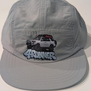 May include: A light gray five-panel cap featuring a graphic of a white SUV with a red cargo carrier and the text "4Runner" in blue. The cap has a curved brim and a structured crown.