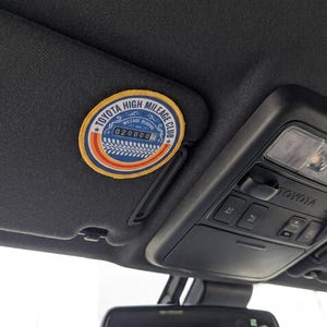 May include: A circular embroidered patch attached to a black vehicle sun visor. The patch features a blue center with the text TOYOTA HIGH MILEAGE CLUB and a mileage reading of 0200000, surrounded by orange and yellow circular borders.