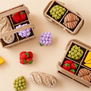 May include: Three small beige plastic baskets containing miniature food items including red strawberries, green grapes, purple grapes, brown bread loaves, croissants, and yellow cheese wedges. Additional loose food items are scattered around.