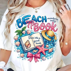 Op de afbeelding: Wit T-shirt met een kleurrijk ontwerp. Het ontwerp bevat de woorden "Beach Book" in blauw en roze, omringd door schelpen, een zeester, een strohoed en een zonnebril. De tekst "Dive into a good book" is ook aanwezig.