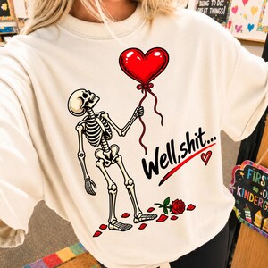 May include: A cream-colored t-shirt featuring a graphic of a skeleton holding a red heart-shaped balloon. The text "Well, shit..." is printed in black script next to the skeleton, accompanied by a single red rose and scattered red rose petals.