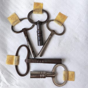 May include: A selection of antique metal keys with diverse handle shapes, including oval and T-forms. Each key is affixed with aged tape. The keys exhibit a worn, vintage aesthetic, indicative of their age and prior use.