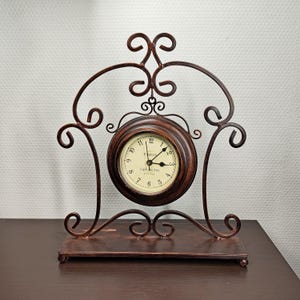 May include: A decorative brown metal clock with a round face and ornate scrollwork. The clock face is cream-coloured with black numerals and hands. The clock reads "Paris Cafe Arts". The clock sits on a rectangular base.