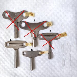 Könnte beinhalten: Eine Sammlung von sechs antiken Metall-Uhrenschlüsseln, jeweils mit einer einzigartigen Kopfform und einem zylindrischen Schaft. Die Schlüssel sind auf einer weißen Oberfläche angeordnet, an einigen sind kleine Klebebänder befestigt.