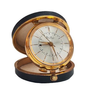 May include: A vintage Belart travel alarm clock in a black leatherette case. The clock has a gold-tone bezel and a silver face with black numerals and hands. The case is open, revealing the clock face and controls. The text "T SWISS MADE T" is visible.