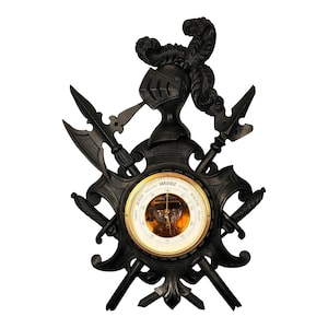 May include: A black, decorative wall-mounted barometer with a knight's helmet and crossed weapons design. The barometer features a gold-colored dial with weather indicators and the word "VARIABLE". The overall design is ornate and detailed.