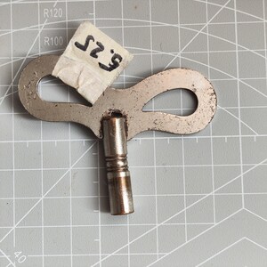 Puede incluir: Una llave de cuerda para reloj de metal plateado con un mango en forma de mariposa y un eje cilíndrico. Un pequeño trozo de cinta adhesiva blanca está pegado al mango, con el texto negro escrito a mano 5.25 en orientación vertical.