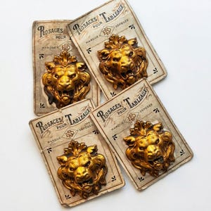 Può includere: Quattro rosette decorative antiche a forma di testa di leone dorata. Ogni rosetta è montata su una carta in stile vintage con testo francese. Le teste di leone hanno criniere dettagliate e bocche aperte.