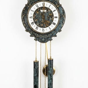 Puede incluir: Un reloj de pared con un marco ornamentado en azul verdoso oscuro y dorado, esfera blanca, números romanos negros y manecillas doradas. Dos pesas cilíndricas con patrón de mármol verde cuelgan de cadenas doradas bajo la esfera del reloj.