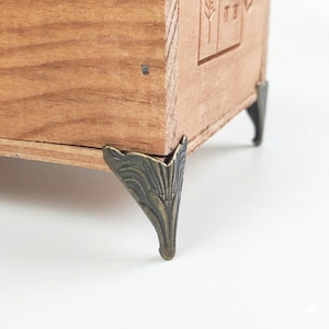 May include: Close-up of a wooden box with decorative metal corner brackets. The box is made of light-colored wood with visible grain. The corner brackets are bronze-colored and feature a leaf-like design. The box appears to be a storage container or decorative item.
