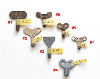 Peut inclure: Un ensemble de sept clés de remontage en métal numérotées de #1 à #7. Chaque clé présente une forme et une taille uniques, certaines marquées par du ruban adhésif jaune indiquant des mesures comme 2,75, 3,0, 2,5 et 4,25. La clé #3 porte l'inscription Joustra Strasbourg France.