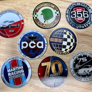May include: A collection of eight circular metal Porsche emblems featuring various designs, including a white RS car, a green helmet, the 356 registry logo, the PCA club logo, a 966 globe, Martini Racing branding, a 70 years badge, and a 75 Jahre emblem.