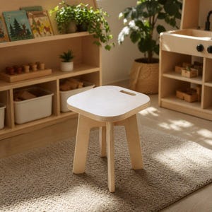 May include: A small, light-colored wooden stool with a square seat and four straight legs. The seat has a small, curved handle. The stool is placed on a neutral-colored rug, with a wooden bookshelf and play kitchen in the background.