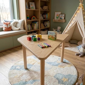 Könnte beinhalten: Ein dreieckiger Tisch aus hellem Holz mit abgerundeten Beinen steht auf einem runden Teppich mit Wolkenmuster. Der Tisch trägt bunte Stapelblöcke, Buntstifte und ein Kinderbuch. Ein Bücherregal und ein Spielzelt befinden sich im Hintergrund.