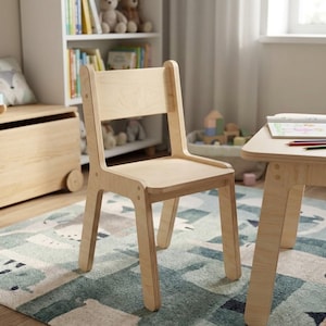 May include: A light-colored wooden chair with a rectangular seat and backrest. The chair has four legs and is designed for children. The chair is part of a set, with a matching table visible in the background.