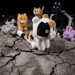 May include: A selection of plush toys in the shape of animals, including cats, a dragon, and a rabbit. The toys are in various colours and have a blocky, pixelated design. The toys are made from soft materials.