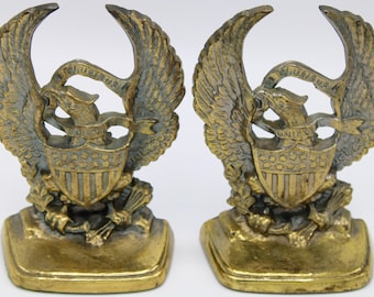 May include: A pair of gold-toned metal bookends featuring an eagle with outstretched wings holding a shield with stars and stripes. The eagle is holding a banner that reads "E Pluribus Unum".