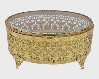 Könnte beinhalten: Eine ovale, goldfarbene Schmuckschatulle mit einem Deckel aus klarem Glas und einem verzierten floralen Filigranmuster. Die Schatulle hat kleine, dekorative Füße und einen Klappdeckel. Die aufwendigen Details und die Vintage-Ästhetik machen sie zu einem charmanten Dekorationsstück.