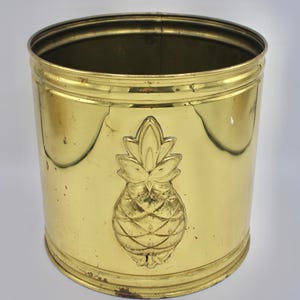 Puede incluir: Un cubo de basura cilíndrico de latón con un emblema de piña. El cubo de basura está hecho de latón y tiene un acabado dorado. El emblema de la piña está en relieve en la parte delantera.
