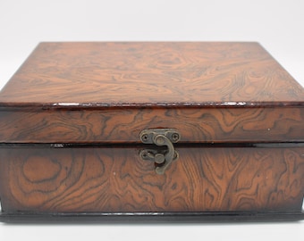 May include: A wooden box with a brown woodgrain finish and a brass latch. The box has a black trim around the edges.