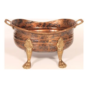 May include: A copper-colored metal decorative bowl with two handles and ornate gold-colored feet. The bowl has a distressed finish and a curved shape, suitable for decorative purposes.