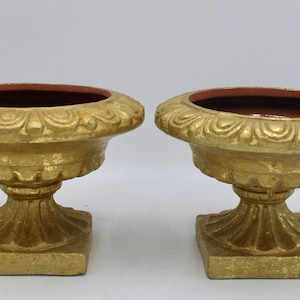 Puede incluir: Dos urnas de cerámica pintadas de oro con bases cuadradas y detalles ornamentados. Las urnas tienen un interior marrón rojizo.